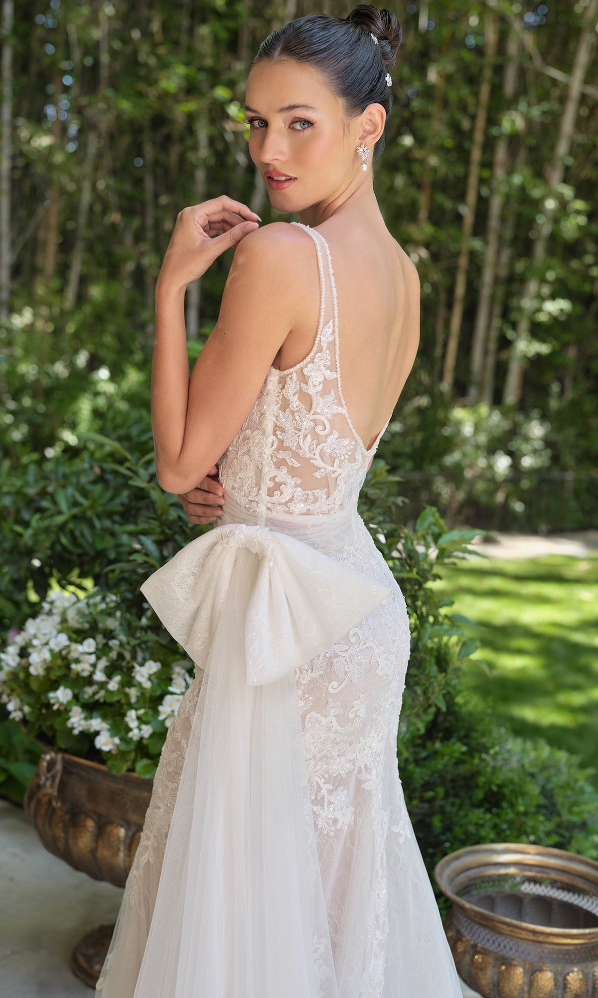 A woman in the Long Wedding Dress: Andrea and Leo WL068 with a lace design, sheer corset bodice, low back, and large bow stands outdoors near greenery, looking over her shoulder.